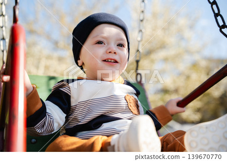 Caucasian baby boy sitting on playground swing in sunny residential park area. Childhood and outdoor lifestyle Caucasian baby boy sitting on playground swing in sunny residential park area. Childhood and outdoor lifestyle 139670770