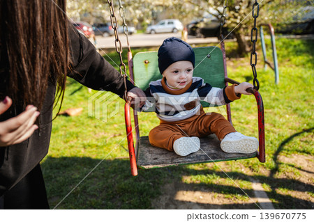 Caucasian baby boy sitting on playground swing with mother nearby in sunny park. Family and childhood lifestyle 139670775