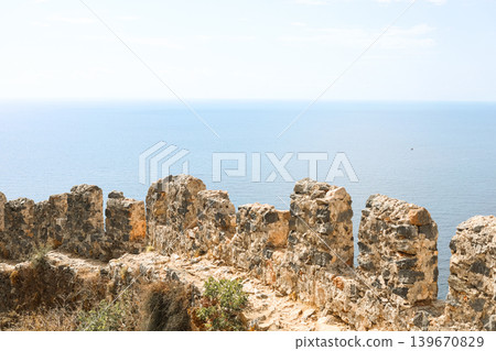 Dilapidated ancient stone wall ruins overlooking calm blue sea horizon Dilapidated ancient stone wall ruins overlooking calm blue sea horizon 139670829