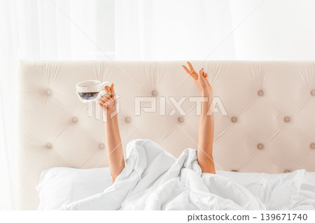 Female hands holding cup and showing peace gesture from under blanket, minimal morning concept with copy space Female hands holding cup and showing peace gesture from under blanket, minimal morning concept with copy space 139671740