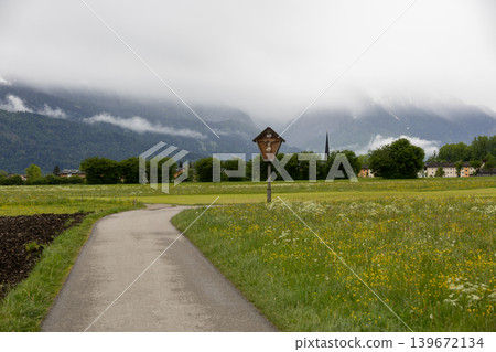 Curving Country Lane Past a Wayside Cross in a Bavarian Alpine Meadow Curving Country Lane Past a Wayside Cross in a Bavarian Alpine Meadow 139672134