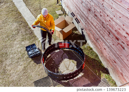 Modular backyard composter with curved plastic walls being constructed by female gardener beside cardboard package. 139673223