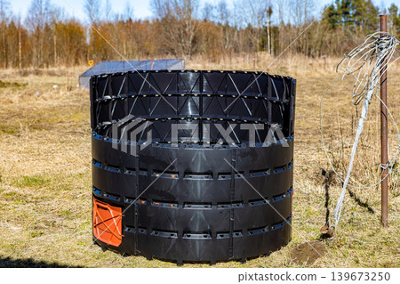 Black circular compost bin with orange door, made of plastic panels, standing outdoors on grass, ready for organic waste processing. 139673250