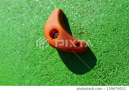 Orange climbing hold mounted on green wall texture, indoor sport equipment, fitness challenge, active lifestyle concept. Orange climbing hold mounted on green wall texture, indoor sport equipment, fitness challenge, active lifestyle concept. 139674031