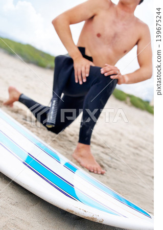 Surfer, man stretching legs and surfboard on beach, fitness and extreme sport, ready for waves with travel and sand. Summer, wet suit and warm up, start surfing and nature with adventure outdoor Surfer, man stretching legs and surfboard on beach, fitness and extreme sport, ready for waves with travel and sand. Summer, wet suit and warm up, start surfing and nature with adventure outdoor 139675244