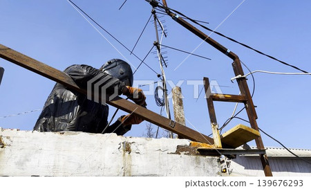 Manual laborer in protective mask welds iron detail at construction. Professional young repairman with welding machine working at the site. Male welder operating with metal on house. Close up Manual laborer in protective mask welds iron detail at construction. Professional young repairman with welding machine working at the site. Male welder operating with metal on house. Close up 139676293