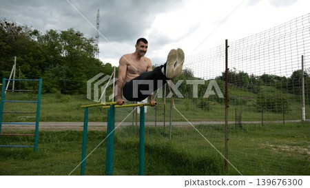 Strong man trains abdominal muscles raising legs on parallel bars at sports ground. Young shirtless sportsman exercising at nature. Sporty athlete working out at nature. Concept of sportive lifestyle Strong man trains abdominal muscles raising legs on parallel bars at sports ground. Young shirtless sportsman exercising at nature. Sporty athlete working out at nature. Concept of sportive lifestyle 139676300