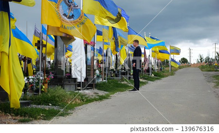 Mourning man standing among countless graves of ukrainian soldiers with national banners pay tribute to fallen family member in Kharkiv. Scene reflect personal sorrow loss caused by russian aggression Mourning man standing among countless graves of ukrainian soldiers with national banners pay tribute to fallen family member in Kharkiv. Scene reflect personal sorrow loss caused by russian aggression 139676316