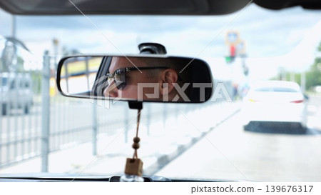 Eyes in sunglasses of male driver reflecting in rearview mirror while riding motor auto. Young guy looking concentrated to urban road during operating vehicle. Confident man driving modern car at city 139676317