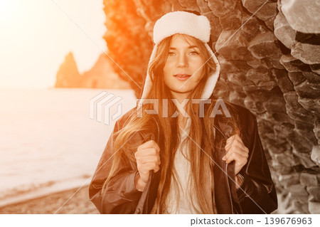 Woman beach sunset Young woman wearing a leather jacket and winter hat poses against a rocky cliff by the sea at sunset Woman beach sunset Young woman wearing a leather jacket and winter hat poses against a rocky cliff by the sea at sunset 139676963