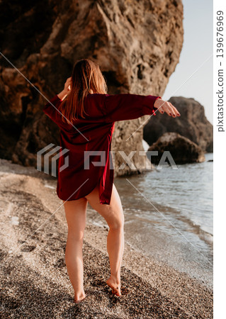 Woman beach vacation standing on a rocky shore wearing a red shirt and facing the ocean during sunrise 139676969