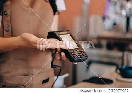 Friendly Asian female barista receives a credit card from a customer for payment. Both women are smiling in a warmly lit coffee shop with a rustic interior. Cashless payments. Small family business 139677052