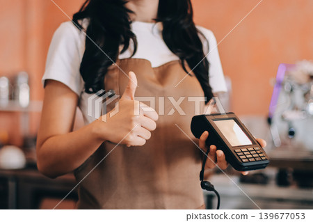 Friendly Asian female barista receives a credit card from a customer for payment. Both women are smiling in a warmly lit coffee shop with a rustic interior. Cashless payments. Small family business Friendly Asian female barista receives a credit card from a customer for payment. Both women are smiling in a warmly lit coffee shop with a rustic interior. Cashless payments. Small family business 139677053