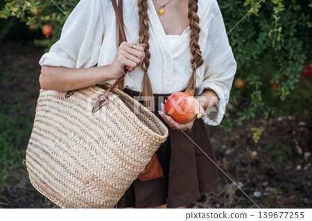 Woman holding apple with woven basket against lush garden background in casual attire Woman holding apple with woven basket against lush garden background in casual attire 139677255
