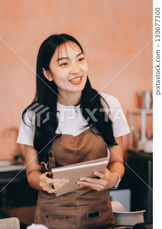 Friendly Asian female barista receives a credit card from a customer for payment. Both women are smiling in a warmly lit coffee shop with a rustic interior. Cashless payments. Small family business Friendly Asian female barista receives a credit card from a customer for payment. Both women are smiling in a warmly lit coffee shop with a rustic interior. Cashless payments. Small family business 139677300