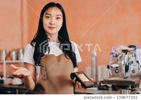 Friendly Asian female barista receives a credit card from a customer for payment. Both women are smiling in a warmly lit coffee shop with a rustic interior. Cashless payments. Small family business 139677302