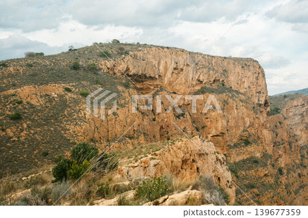 Dramatic rocky cliffs with lush vegetation under cloudy sky in canyon landscape Dramatic rocky cliffs with lush vegetation under cloudy sky in canyon landscape 139677359