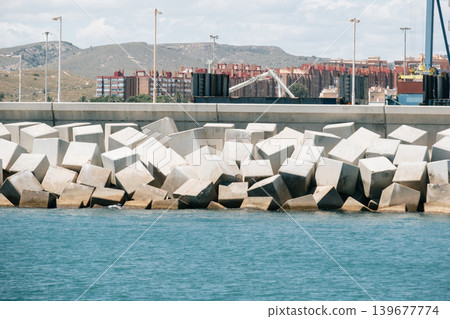 Coastal breakwater with concrete blocks near urban waterfront and hills in the background Coastal breakwater with concrete blocks near urban waterfront and hills in the background 139677774