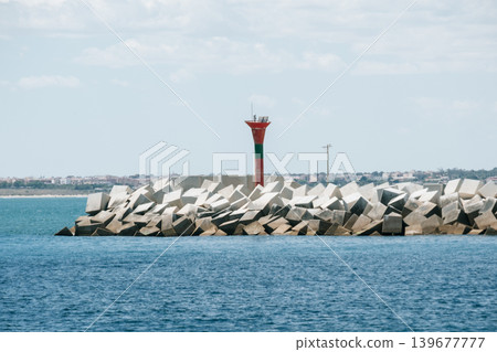 Coastal breakwater with red lighthouse tower and geometric concrete blocks under blue sky Coastal breakwater with red lighthouse tower and geometric concrete blocks under blue sky 139677777