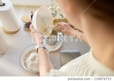 Woman scrapes flour from bowl into sifter beside glass bowl with batter Woman scrapes flour from bowl into sifter beside glass bowl with batter 139678753