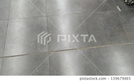 Dark gray floor tiles feature a subtle textured pattern. Bright indoor lighting reflects off the smooth surface, creating a soft silver glow. Straight grout lines divide the large square panels. The Dark gray floor tiles feature a subtle textured pattern. Bright indoor lighting reflects off the smooth surface, creating a soft silver glow. Straight grout lines divide the large square panels. The 139679865