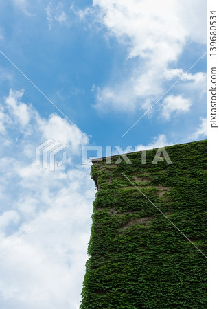 A low-angle photograph of a building with ivy-covered exterior walls, blue sky, and clouds, showcasing the green architecture. A low-angle photograph of a building with ivy-covered exterior walls, blue sky, and clouds, showcasing the green architecture. 139680534