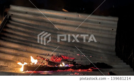 A fire blazes on a corrugated metal roof, casting a fiery glow and revealing glowing embers. The long exposure captures the motion of the flames and rising smoke against the dark night. A fire blazes on a corrugated metal roof, casting a fiery glow and revealing glowing embers. The long exposure captures the motion of the flames and rising smoke against the dark night. 139681214