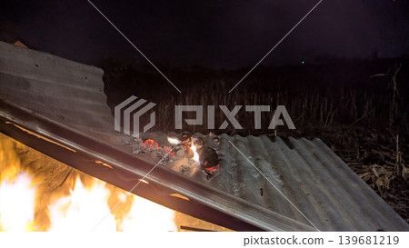 A wide-angle long exposure shot captures a fire on a corrugated metal roof. The bright flames and glowing embers are seen against the dark night sky with a field in the background. 139681219