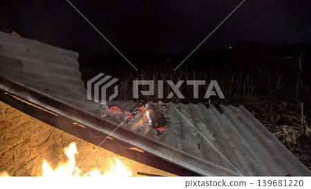 A wide-angle long exposure captures a fire on a corrugated metal roof. The bright flames and glowing embers are seen against the dark night sky with a field and brick wall in the background. A wide-angle long exposure captures a fire on a corrugated metal roof. The bright flames and glowing embers are seen against the dark night sky with a field and brick wall in the background. 139681220
