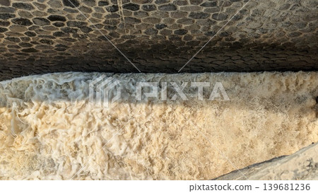 A view of foaming, turbulent water with a yellowish-brown tint rushing under a stone barrier or bridge from above the water appears frothy and agitated, contrasting with the dark, textured stone A view of foaming, turbulent water with a yellowish-brown tint rushing under a stone barrier or bridge from above the water appears frothy and agitated, contrasting with the dark, textured stone 139681236