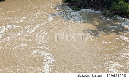 A murky brown river flows rapidly past a vegetated bank where debris is caught. The water is churning with white foam and reflections of overhead shadows. It appears muddy after heavy rain. A murky brown river flows rapidly past a vegetated bank where debris is caught. The water is churning with white foam and reflections of overhead shadows. It appears muddy after heavy rain. 139681238