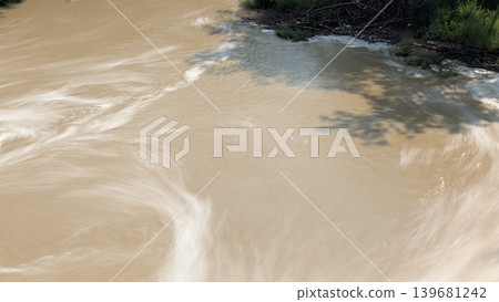 An artistic long exposure of a muddy brown river, showing silky white swirls of moving water, with a dark bank of greenery, foam, debris on the top edge, a shadow of an overhead object. An artistic long exposure of a muddy brown river, showing silky white swirls of moving water, with a dark bank of greenery, foam, debris on the top edge, a shadow of an overhead object. 139681242