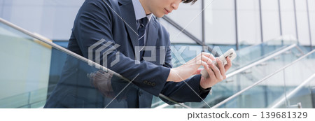 一位商人站在建築物的室外樓梯上,用智慧型手機敲擊地面,橫幅上顯示著他嚴肅的表情。 一位商人站在建築物的室外樓梯上,用智慧型手機敲擊地面,橫幅上顯示著他嚴肅的表情。 139681329