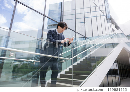 一位商人站在建築物的室外樓梯上,用智慧型手機敲擊螢幕——一個冷靜、英俊的男人,正在執行銷售任務。 一位商人站在建築物的室外樓梯上,用智慧型手機敲擊螢幕——一個冷靜、英俊的男人,正在執行銷售任務。 139681333