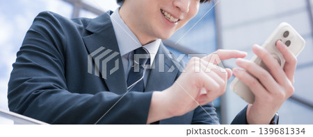一位商人站在建築物的室外樓梯上,用智慧型手機敲擊螢幕;一位英俊瀟灑的男士拿著一部沒有面孔的智慧型手機。 一位商人站在建築物的室外樓梯上,用智慧型手機敲擊螢幕;一位英俊瀟灑的男士拿著一部沒有面孔的智慧型手機。 139681334