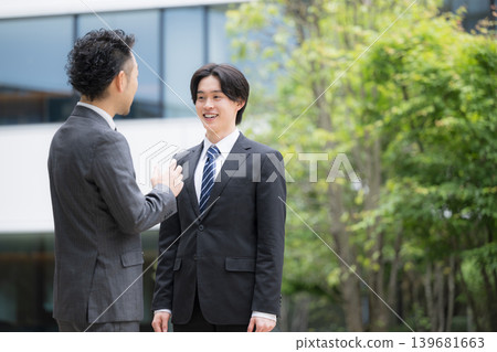 A trustworthy sales team in suits communicates in front of a building outdoors; a young subordinate (newcomer) and their superior are having a casual chat. A trustworthy sales team in suits communicates in front of a building outdoors; a young subordinate (newcomer) and their superior are having a casual chat. 139681663