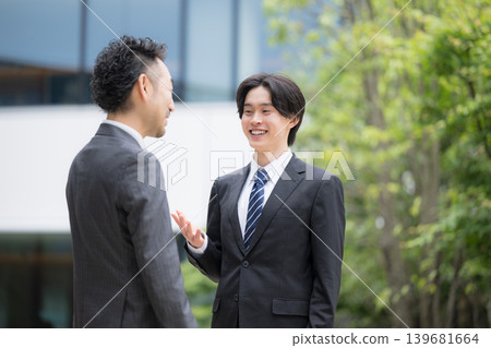 A trustworthy sales team in suits communicates in front of a building outdoors; a young subordinate (newcomer) and their superior are having a casual chat. A trustworthy sales team in suits communicates in front of a building outdoors; a young subordinate (newcomer) and their superior are having a casual chat. 139681664
