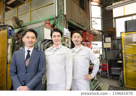 A team of three salesmen in work clothes and suits working on the factory floor of a manufacturing plant; smiling directly at the camera, representing a small business. A team of three salesmen in work clothes and suits working on the factory floor of a manufacturing plant; smiling directly at the camera, representing a small business. 139681670