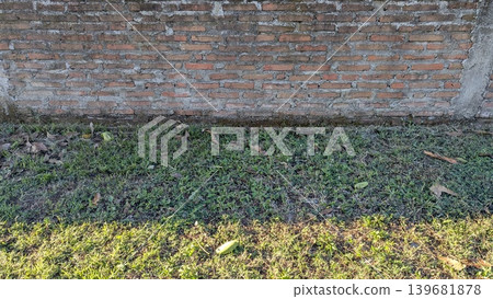 A rustic brick wall sits above a patch of green and brown grass with some fallen leaves. A sliver of sunlight illuminates the foreground creating a sharp contrast. A rustic brick wall sits above a patch of green and brown grass with some fallen leaves. A sliver of sunlight illuminates the foreground creating a sharp contrast. 139681878