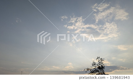 A serene twilight sky displaying crepuscular rays fanning out from behind a silhouetted treetop at the bottom right. Delicate white clouds drift across the soft blue and gray gradient. A serene twilight sky displaying crepuscular rays fanning out from behind a silhouetted treetop at the bottom right. Delicate white clouds drift across the soft blue and gray gradient. 139681943