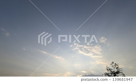 A calm evening sky featuring a subtle gradient from deep blue to soft white and gold at the horizon. Scattered white clouds catch the light, and crepuscular rays fan out from below the frame. 139681947