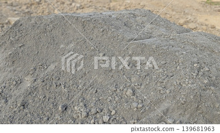 A close-up of a large pile of dark gray fine aggregate or volcanic sand, showing its rough texture and particle variation, set against a blurred background of lighter-colored earth. A close-up of a large pile of dark gray fine aggregate or volcanic sand, showing its rough texture and particle variation, set against a blurred background of lighter-colored earth. 139681963