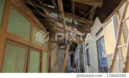 Old wooden roof beams collapse between two narrow village houses. Weathered green doors, peeling paint, bamboo ladders create a scene of decay. Sunlight hits the white walls, casting deep shadows over 139682686