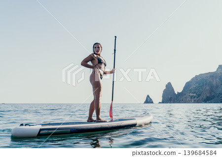 Woman Paddleboarding Ocean Water Sports - A woman in a swimsuit is standing on a paddleboard in the middle of the ocean with mountains in the background. Woman Paddleboarding Ocean Water Sports - A woman in a swimsuit is standing on a paddleboard in the middle of the ocean with mountains in the background. 139684584