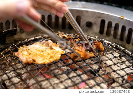 Close up of meat grilling on a charcoal barbecue grill with hands using metal chopsticks to turn the food. Traditional yakiniku or Korean BBQ dining experience with glowing embers and smoke. Close up of meat grilling on a charcoal barbecue grill with hands using metal chopsticks to turn the food. Traditional yakiniku or Korean BBQ dining experience with glowing embers and smoke. 139687468