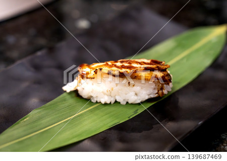 Single piece of grilled unagi sushi nigiri served on a fresh green leaf over a black plate. Close-up food photography showcasing the glazed texture and white rice. High-end restaurant presentation. Single piece of grilled unagi sushi nigiri served on a fresh green leaf over a black plate. Close-up food photography showcasing the glazed texture and white rice. High-end restaurant presentation. 139687469