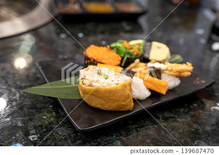 Assorted sushi platter on a black plate featuring inari with tuna, crab stick nigiri, and seaweed salad. Close-up of fresh Japanese cuisine served on a dark stone table in a restaurant setting. 139687470