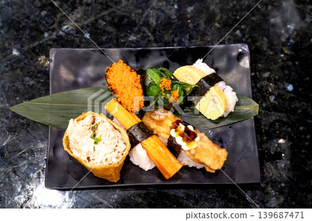 Assorted sushi platter on a black ceramic plate over dark marble background. Includes inari, crab stick nigiri, tamago, fried topping, seaweed salad, and fish roe. Traditional Japanese food garnish. Assorted sushi platter on a black ceramic plate over dark marble background. Includes inari, crab stick nigiri, tamago, fried topping, seaweed salad, and fish roe. Traditional Japanese food garnish. 139687471