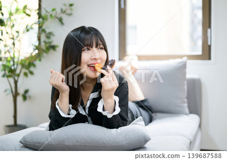 Japanese woman relaxing in bed while eating a donut. Japanese woman relaxing in bed while eating a donut. 139687588