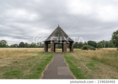 Open brick pavilion standing beside grassy fields under dramatic cloudy skies. Peaceful countryside scenery creates a calm outdoor landscape atmosphere. Open brick pavilion standing beside grassy fields under dramatic cloudy skies. Peaceful countryside scenery creates a calm outdoor landscape atmosphere. 139688468